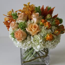 Short arrangement of peach roses, orange blooms, and white hydrangeas in a clear glass cube vase