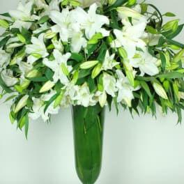 Tall arrangement of white lilies and buds in a slender green glass vase