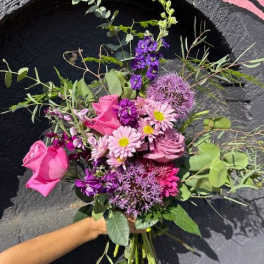Hand-tied bouquet of pink and purple flowers with long stems held against a dark textured wall