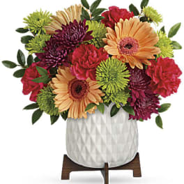Short mixed bouquet of peach gerberas, red carnations, and green mums in a white pot on a wooden stand