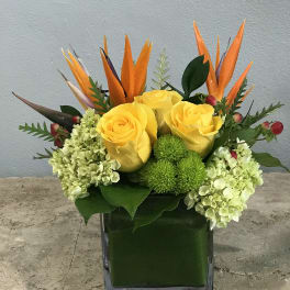 Yellow roses and green hydrangeas in a square glass vase with orange tropical accents