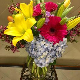 Bright mixed bouquet with yellow lilies, pink gerberas, and blue hydrangeas in a glass vase