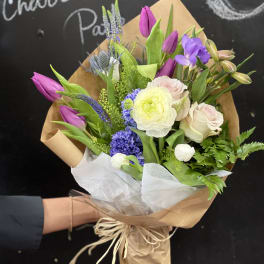 Hand-tied bouquet of purple tulips, pale roses, and mixed blooms in brown paper