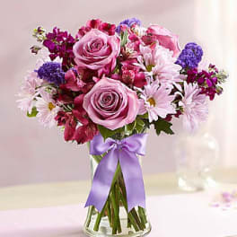 Pink roses and purple flowers in a clear glass vase with a lavender ribbon