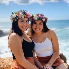 Two people wearing colorful floral crowns sitting by the ocean on a sunny day