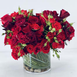 Compact arrangement of red roses in a clear glass cylinder vase with visible spiraled stems and stones.