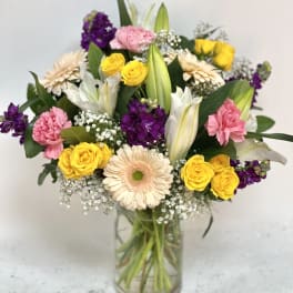 Mixed bouquet of yellow roses, pink carnations, lilies, and daisies in a glass vase