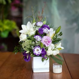 Purple and white floral arrangement in a white square vase beside a small candle