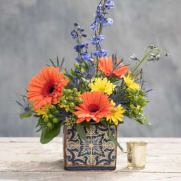Orange and yellow flowers in a patterned square vase with a small candle holder beside it