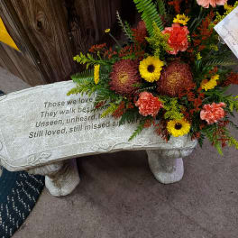 Sympathy spray with yellow daisies and peach carnations on a memorial bench