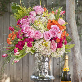 Large pink and orange floral arrangement in a glass vase beside a champagne bottle