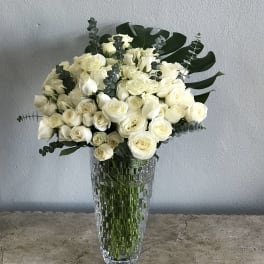 White rose bouquet in a tall clear glass vase with dark green foliage