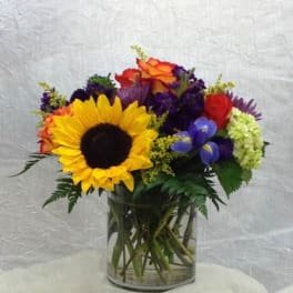 Mixed bouquet with a sunflower, roses, and purple flowers in a glass vase