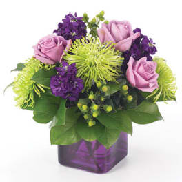 Lavender roses, green spider mums, and purple flowers in a purple glass cube vase