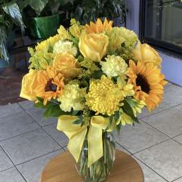 Yellow bouquet of roses, sunflowers, and mums in a glass vase with a ribbon