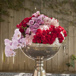 Pink, red, and lavender roses arranged in a silver urn with orchids draping over the side