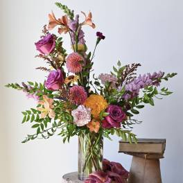 Tall mixed arrangement of purple, pink, and orange flowers in a clear glass vase beside stacked books.