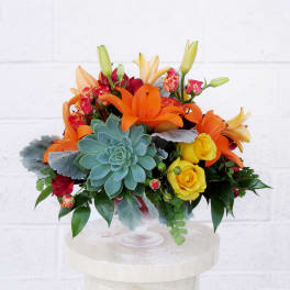 Bright bouquet of orange lilies, yellow roses, and a blue-green succulent in a glass vase.