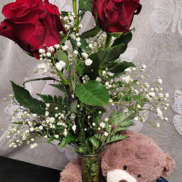 Three red roses in a glass vase with a teddy bear beside it