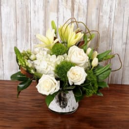 Compact arrangement of white roses, lilies, hydrangeas, and green blooms in a rustic glass vase.