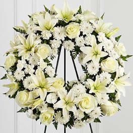White floral wreath on a black stand with lilies and roses