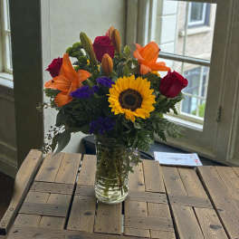 Bouquet of sunflowers, red roses, orange lilies, and purple filler in a glass vase