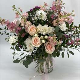 Tall arrangement of peach roses, white mums, and pink blooms in a glass vase with trailing ribbons
