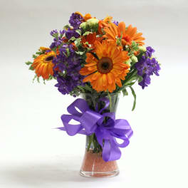 Orange and purple mixed bouquet in a clear glass vase with a purple ribbon
