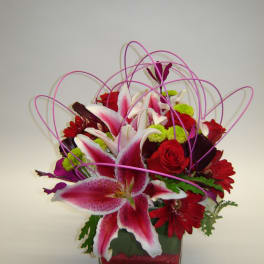Pink lilies and red roses in a square glass vase with looping purple accents