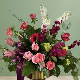 Pink and red mixed flower arrangement in a gold vase with a purple ribbon