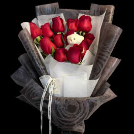 Hand-tied bouquet of red roses wrapped in dark paper with a small white teddy bear.