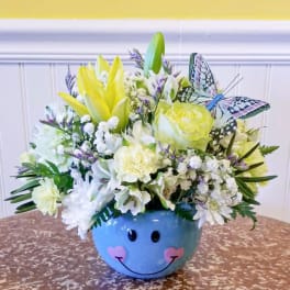 Yellow and white flower arrangement in a blue smiley-face bowl with a butterfly decoration