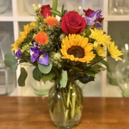 Mixed bouquet of sunflowers, red roses, and purple flowers in a glass vase