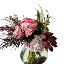 Pink roses, hydrangeas, and dark tulips arranged in a round glass vase.