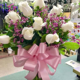 White roses and purple filler flowers in a glass vase with a pink bow