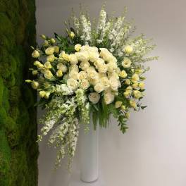 Tall grand arrangement of ivory roses and white flowers in a white pedestal vase against a moss wall