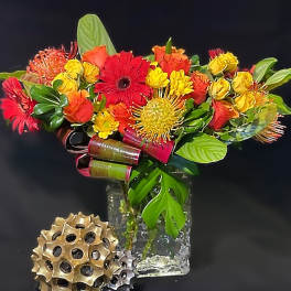 Bright red, orange, and yellow flower arrangement with tropical accents in a clear glass cube vase