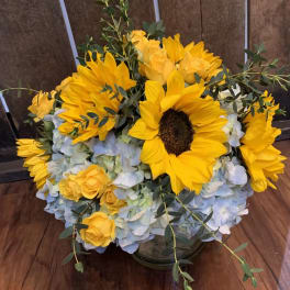 Yellow sunflowers and roses in a low glass vase with pale blue hydrangeas.