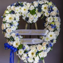 Heart-shaped floral wreath with white flowers and blue accents on a stand