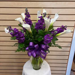 Purple flowers and white calla lilies in a glass vase with a purple ribbon