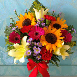 Mixed bouquet with sunflowers, lilies, gerbera daisies, and a red ribbon in a glass vase