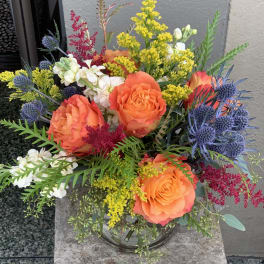 Bouquet of orange roses, white blooms, and blue thistles in a glass vase