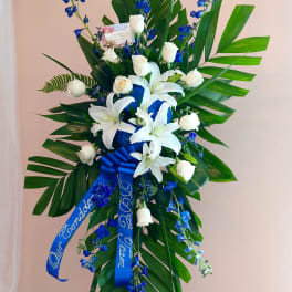 Tall standing floral spray with white lilies, white roses, and blue flowers on an easel.
