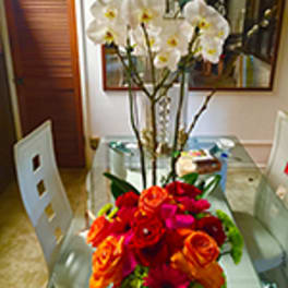 Tall white orchids with a bright cluster of red and orange roses in a clear vase on a glass table