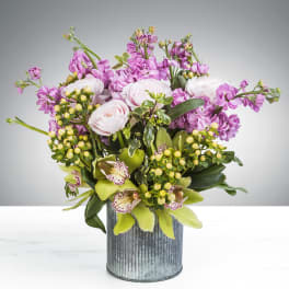 Lavender and pale blooms with green orchids and berries arranged in a ribbed metal cylinder vase.