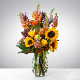 Tall arrangement of sunflowers, yellow roses, and orange snapdragons in a clear glass vase