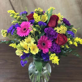 Mixed bouquet of red roses, pink gerbera daisies, and yellow and purple flowers in a glass vase