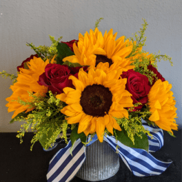 Low arrangement of bright sunflowers and red roses in a metal vase with a striped blue ribbon.