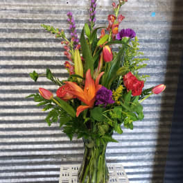 Tall mixed bouquet in a clear glass vase with lilies, tulips, and purple spikes