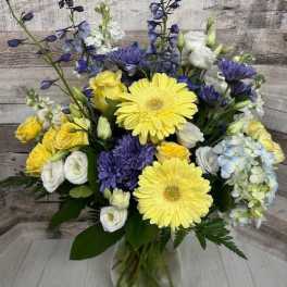 Bouquet of yellow gerbera daisies, blue flowers, and white blooms in a glass vase